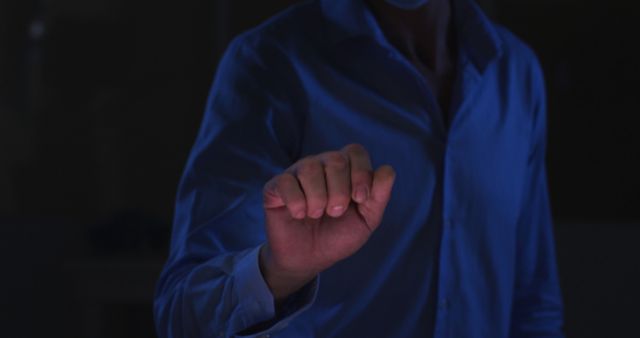 A man in a blue shirt is gesturing with his hand in a dark room. This image can be used to represent themes of technology, futuristic interaction, communication, and nighttime activities. Perfect for use in technology-related promotions, articles about cutting-edge innovations, or slideshows depicting interactive environments.
