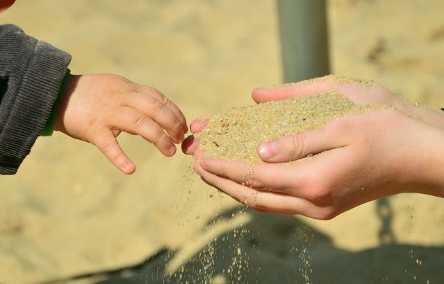 A child's hand reaches towards sand held by an adult hand, reflecting playful curiosity and bonding on a beach. Useful for themes of childhood, parenting, and outdoor activities. Suitable for articles, blogs, or advertisements focusing on family time, play, or beach activities.