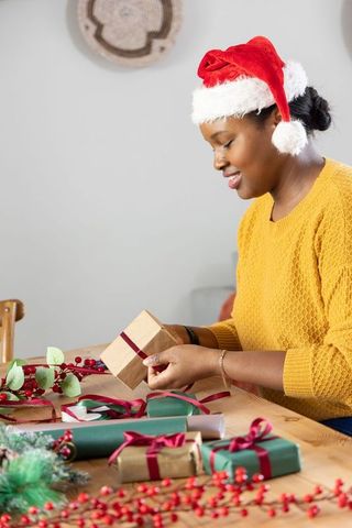 African american woman wrapping christmas gifts with red ribbon wearing santa hat