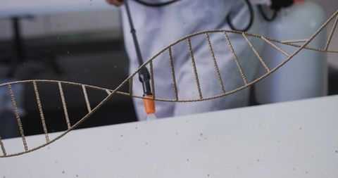 Rope dna helix suspended over workbench, technician spraying orange nozzle conserving artwork