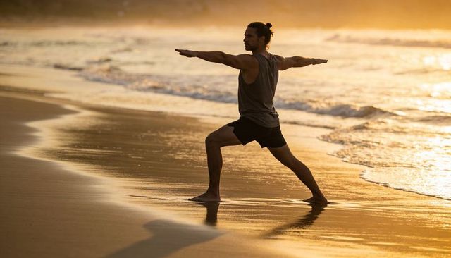 Male practicing warrior ii yoga pose on golden beach at sunset with serene ocean reflection