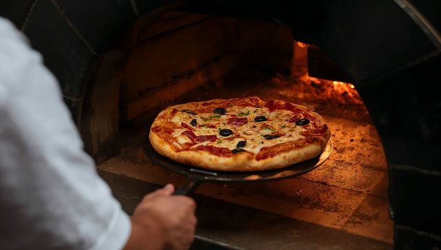 Chef sliding wood-fired pizza from brick oven with bubbling cheese, pepperoni, olives