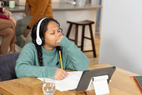 Tired girl wearing headphones yawning while studying at kitchen table with tablet and notebook