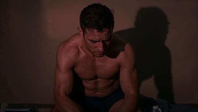 Muscular man resting after intense workout, leaning forward and contemplating in low light