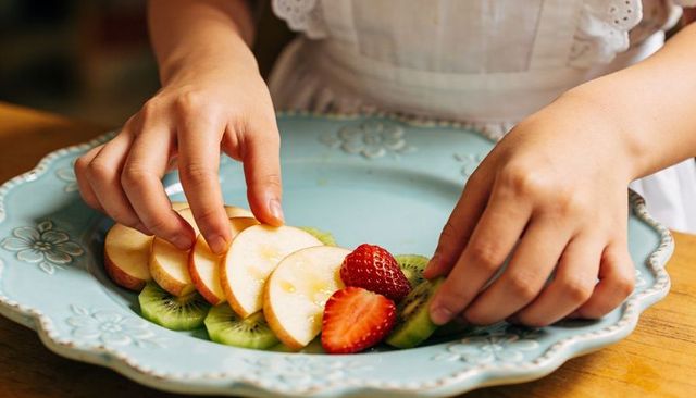 Child arranging sliced apple kiwi strawberry on embossed turquoise ceramic plate close-up