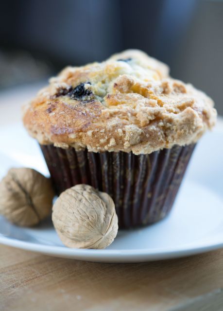A close-up of a freshly baked muffin with a crumbly top placed on a white plate, accompanied by two whole walnuts. Ideal for use in marketing bakery products, showcasing breakfast or dessert items, or promoting homemade baked goods.