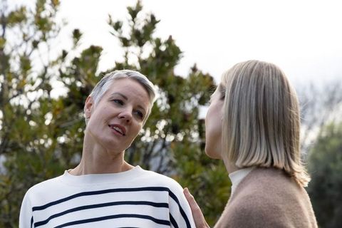Senior woman and friend chatting outdoors by evergreen shrubs, relaxed mature companionship