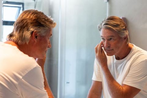 Mature man checking under-eye skin and grooming at bathroom mirror, self-care routine