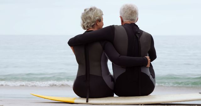 Senior couple embracing while sitting on a surfboard on the beach, looking out at the ocean. Ideal for themes related to elderly companionship, love and adventure, retirement lifestyle, outdoor activities, and active aging.