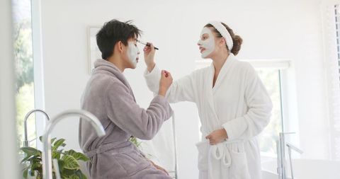 Couple applying facial masks in bright bathroom wearing plush bathrobes, cozy home spa