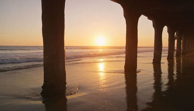 Casting golden sunset reflections through pier columns along calm ocean shoreline