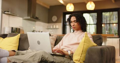 Non-binary person working on laptop on cozy gray sofa with yellow cushions in modern open-plan home