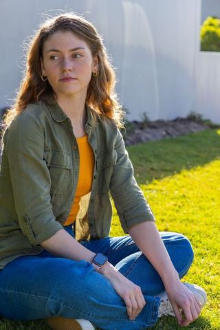 Young woman sitting on sunlit lawn looking thoughtful wearing olive jacket and smartwatch