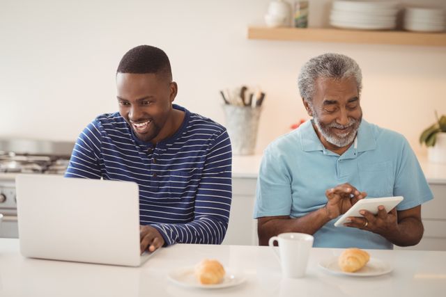 Father and son are using laptop and tablet in kitchen, experiencing joy and connectivity. Perfect for illustrating family bonding, home life, and the use of technology across generations. Suitable for promoting digital products, family activities, and modern lifestyle concepts.