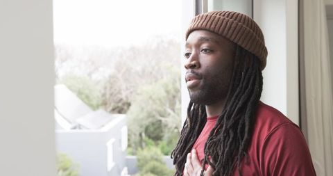 African American man gazing out window wearing brown beanie and dreadlocks, contemplative home momen