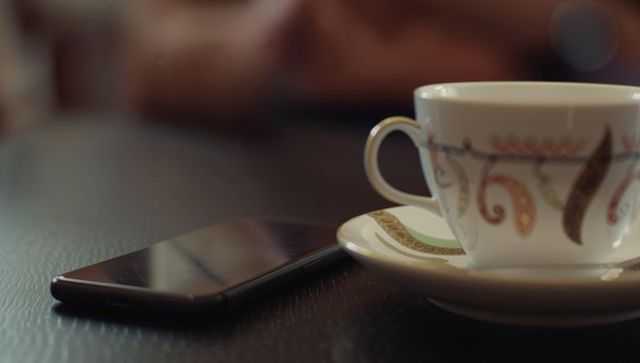 Porcelain teacup resting on paisley saucer next to smartphone on dark table