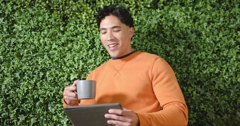 Asian man sipping from gray mug while using tablet by lush green hedge, casual lifestyle