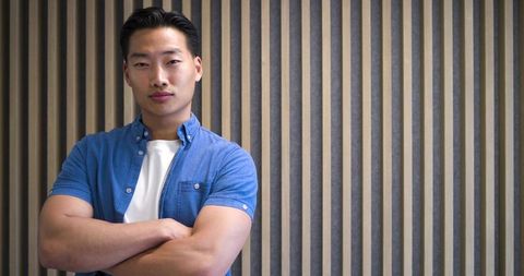 Confident young asian man standing with arms crossed facing camera against modern slat wall