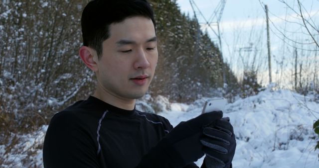 An Asian man is checking his smartphone during a break in a snowy outdoor environment, with copy space. He appears to be engaged in his device, tracking his fitness progress or navigating a route.