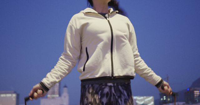 Woman exercising by jumping rope at dusk in an urban area with cityscape background. Dressed in sportswear and deeply concentrated on her workout. Ideal for illustrating fitness routines, outdoor activities, and healthy lifestyle concepts in urban settings.