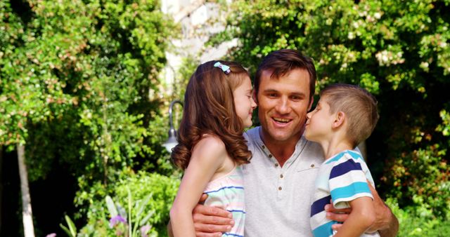 A middle-aged Caucasian man enjoys a sunny day outdoors with two children, a boy and a girl, showing affection with kisses on the cheek. Their joyful interaction captures a moment of family bonding and love in a natural setting.