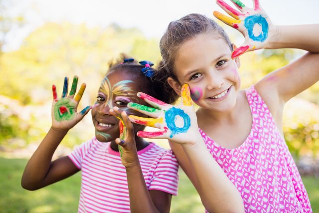 Two young girls with face paint and colorful hands are smiling and enjoying their time in a park. This image is perfect for promoting children's activities, summer camps, art classes, and outdoor events. It highlights themes of friendship, creativity, and the joy of childhood.