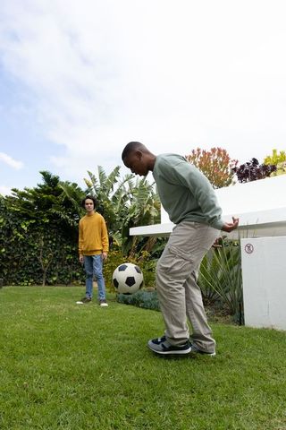 Friends playing soccer on backyard lawn with tropical garden casual clothing and bench