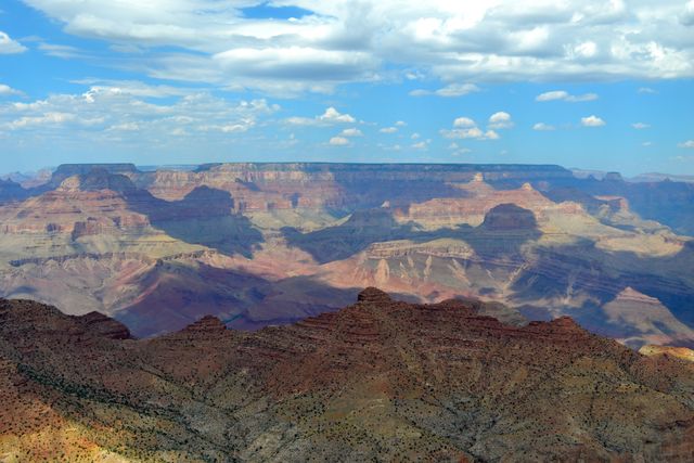 Panoramic view showcasing the vast and stunning landscape of the Grand Canyon under a partially cloudy sky. Presence of dramatic shadows and light emphasizes the unique rock formations and varied topography. Ideal for use in travel and adventure promotions, outdoor magazines, and educational materials about natural wonders and geology.