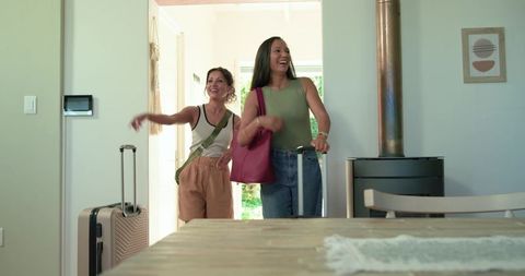 Diverse female friends arriving at bright home carrying luggage and laughing