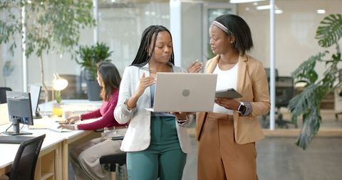 Two african american professional women collaborating over laptop in modern bright office