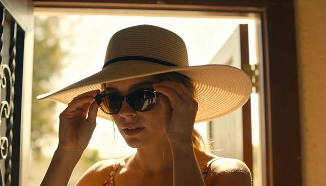 Woman adjusting sunglasses wearing wide-brim straw hat at golden hour doorway