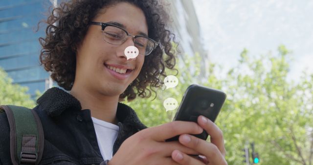 Young man with curly hair and glasses smiling while chatting on smartphone in an urban outdoor setting. Perfect for illustrating themes of modern communication, social media, technology use, and outdoor city life. Ideal for websites, blogs, or advertisements focusing on youth culture, digital trends, or tech devices.