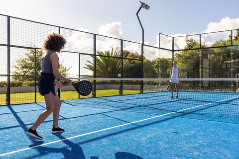 Women playing padel on bright blue court swinging paddle under sunny sky outdoor doubles match