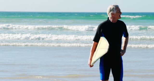 Senior man standing on beach with surfboard while wearing wetsuit and looking at ocean waves. Perfect for illustrating active lifestyles in retirement, leisure activities, and coastal tourism. Could be used for fitness, travel, or retirement-related content.