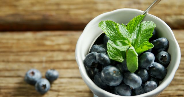 A bowl of fresh blueberries topped with a mint leaf is presented on a rustic wooden surface, with copy space. Blueberries are known for their antioxidant properties and are a popular choice for a healthy snack or dessert ingredient.