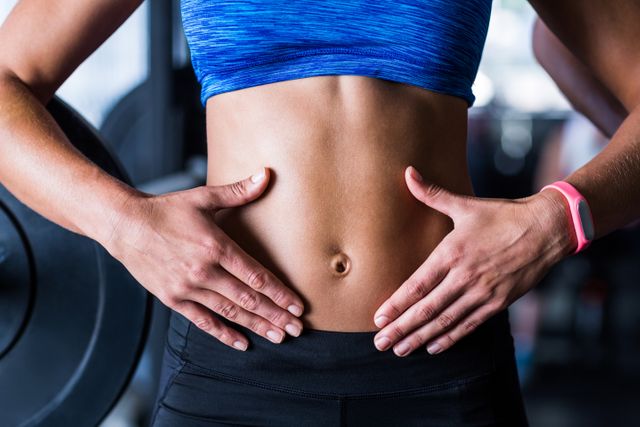 Midsection of female athlete touching her belly while standing in gym. Ideal for fitness blogs, workout programs, health and wellness articles, sportswear advertisements, and gym promotions.