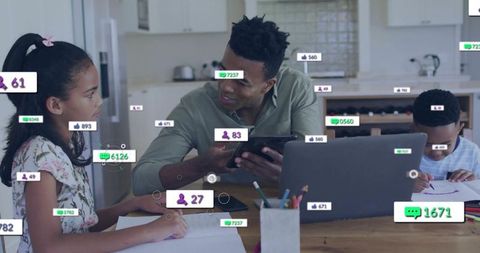 Father helping children with digital learning at kitchen table using tablet and laptop