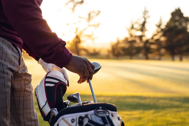 Midsection of African American young man organizing golf clubs in bag at golf course during sunset. Ideal for use in advertisements, sports promotions, leisure activity content, and lifestyle blogs focusing on outdoor activities and golfing.