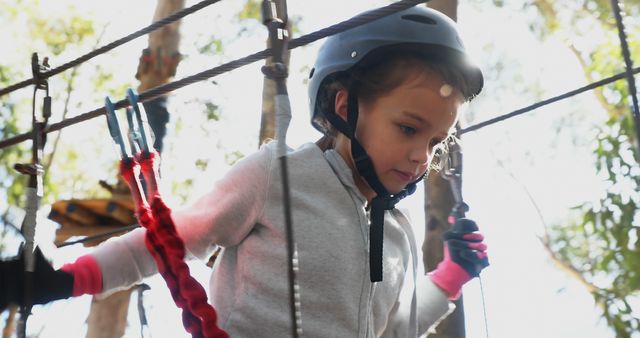 A young Caucasian girl is navigating an outdoor adventure course, wearing a safety helmet and focused on her next move. Her determination and the challenges of the course emphasize the importance of physical activity and confidence-building in childhood.