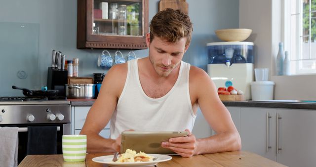 A young man in casual attire using a digital tablet while having breakfast in a modern kitchen. Ideal for themes related to morning routines, technology in everyday life, work from home, student life, and digital devices.