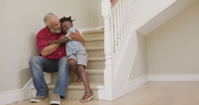 Elderly man sitting on staircase holding and comforting a young child in a brightly lit home, showcasing family bonding time and emotional support. The image is perfect for projects focusing on multigenerational relationships, affection, family life, and the warmth of home. Useful for websites, advertisements, and articles about family dynamics, emotional well-being, and indoor activities.