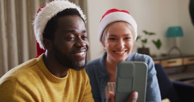 Multicultural couple wears matching Santa hats and smiles while taking a selfie with a smartphone in a cozy indoor setting. Ideal for holiday-themed advertisements, social media posts, and projects highlighting festive joy and togetherness.