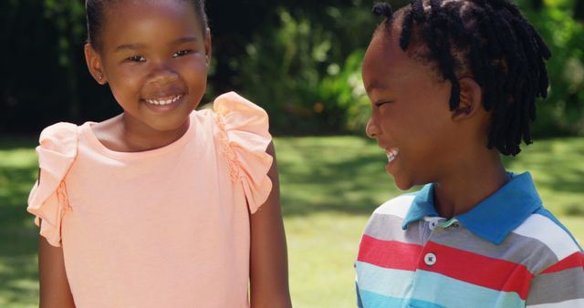 Two African American children are seen smiling outdoors in a park setting with greenery in the background. The girl is wearing a pink dress and the boy is wearing a striped polo shirt. This image conveys happiness and can be used for promoting children's activities, summer fun, family bonding, or community and friendship themes.