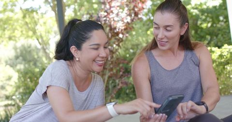 Two women sharing smartphone on park bench, smiling and pointing, wearing fitness trackers