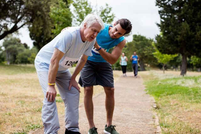 Senior man looking exhausted after a race in a park, receiving support from a younger man. Ideal for use in articles or advertisements about senior fitness, health and wellness, outdoor activities, and the importance of support and encouragement in physical activities.