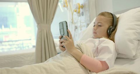 Child patient lying in hospital bed using smartphone and headphones near infusion pumps