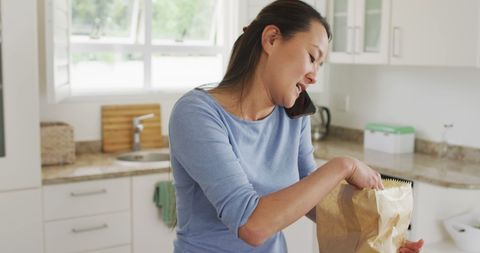 Asian Woman Multitasking Using Phone and Unpacking Groceries in Modern Kitchen