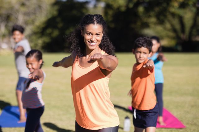 Female yoga trainer leading a group of children in Virabhadrasana II pose in a park. Ideal for promoting outdoor fitness, family activities, wellness programs, and children's exercise classes.