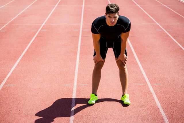 Young male athlete taking a break on a running track during a sunny day. Ideal for use in fitness, sports training, and athletic performance content. Can be used to illustrate themes of endurance, determination, and the physical demands of competitive sports.