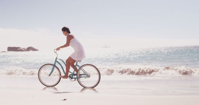 A young Caucasian woman enjoys a leisurely bike ride along the sandy shore of a beach, with copy space. Her white dress and the clear blue sky create a serene and refreshing summer atmosphere.
